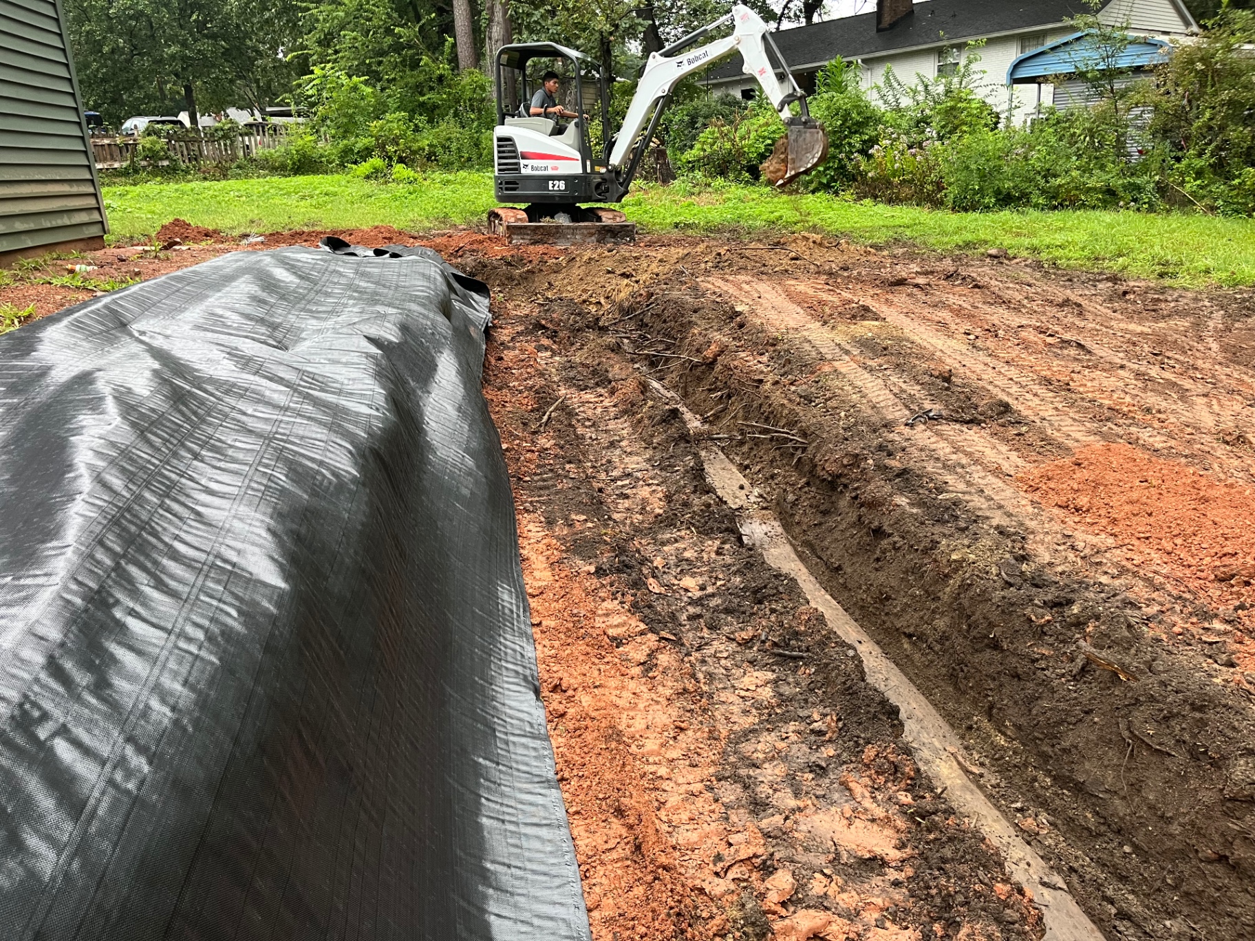 Bobcat E26 mini excavator digging a pipe trench in red clay beside a roll of filter fabric, Lincolnton, NC
